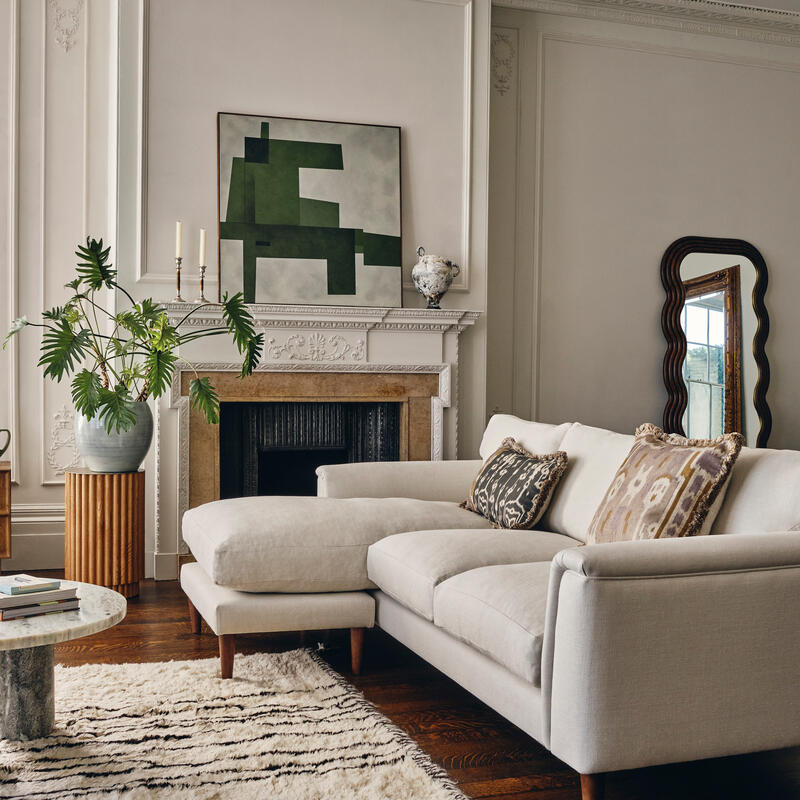 A light grey sectional sofa with patterned cushions rests on a white, textured rug in an elegantly decorated living room featuring a fireplace, large abstract painting, potted plant, and mirror.