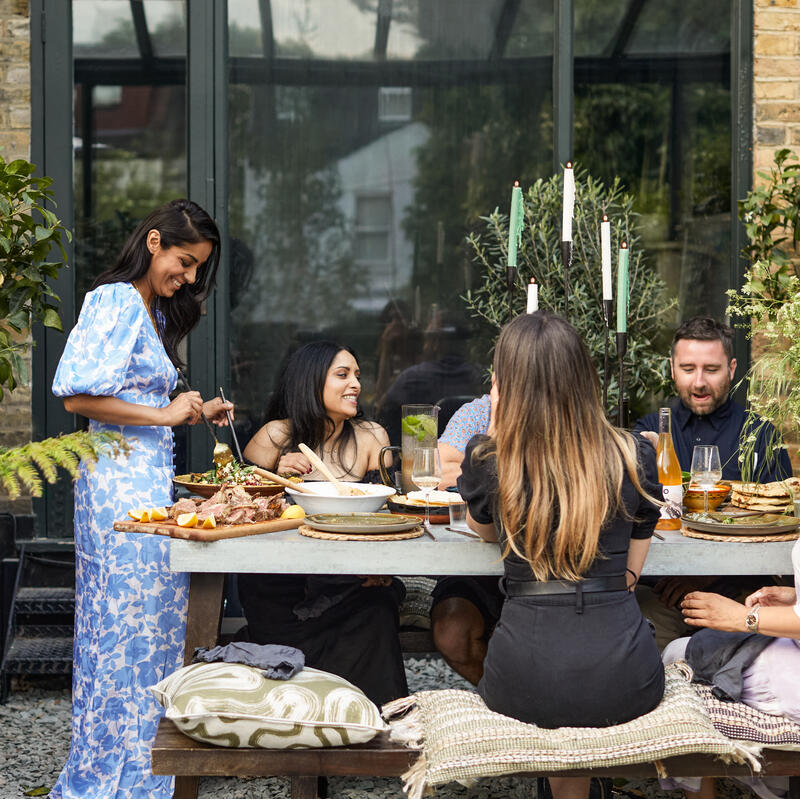 People dining together, enjoying food and drinks around a table outdoors amidst a lush, garden environment with a brick wall and large glass windows in the background.