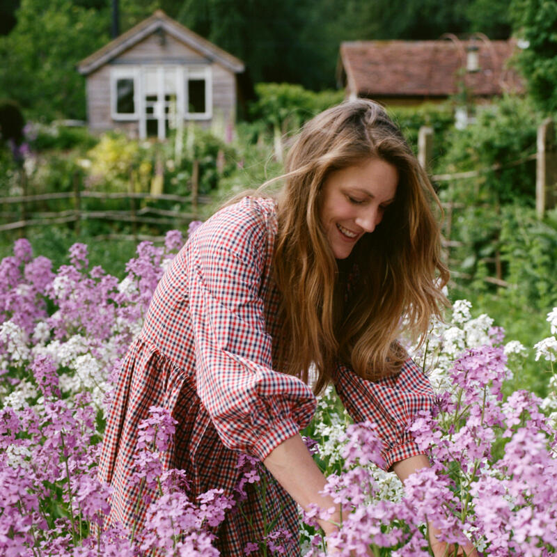 Woman in a plaid dress gathers pink and white flowers in a thriving garden with a small wooden house in the background.