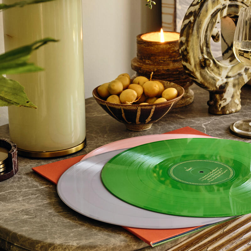 A green and white vinyl records rest on a marble table alongside a table lamp, potted plant, candles, bowl of small potatoes, and wine glass in a cozy, decorated living space.