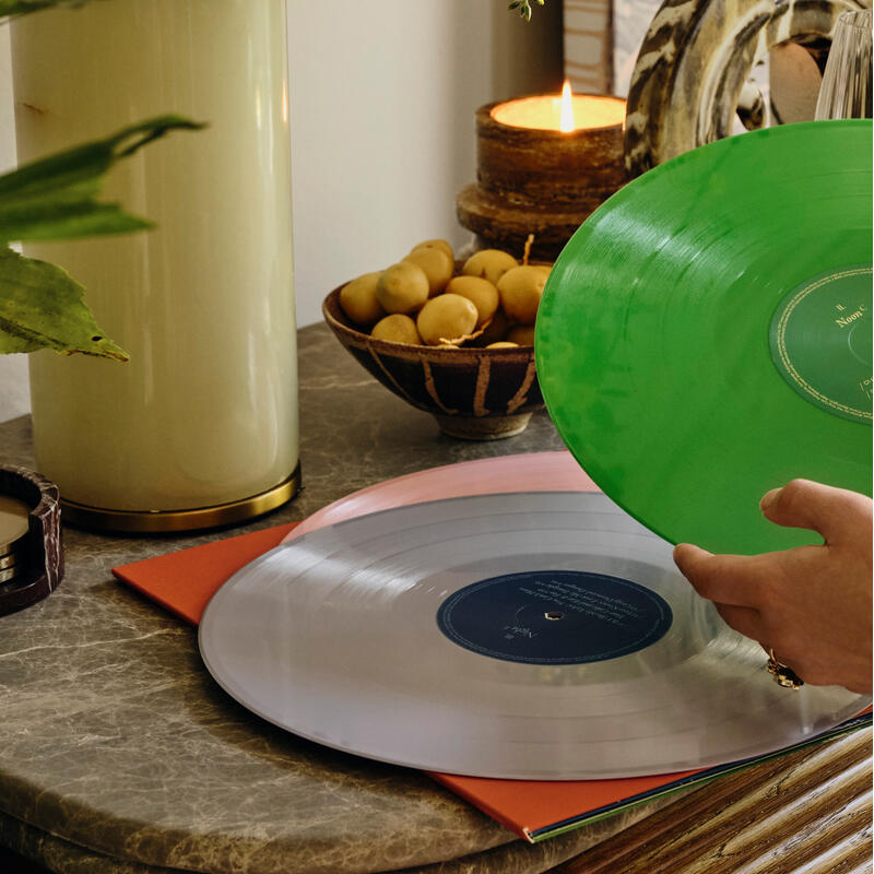 A person holds a green vinyl record over a table with a pink vinyl record, a bowl of yellow fruits, a lit candle, a plant, and a lamp in a cozy setting.