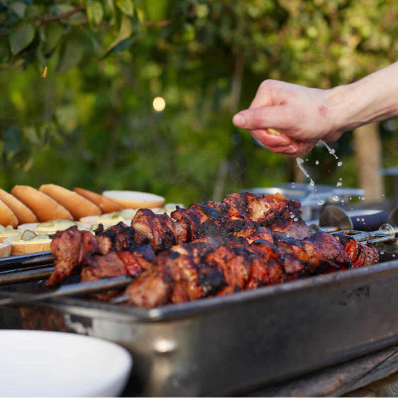 Grilled skewers of meat char slightly, with someone sprinkling liquid over them; hot dog buns are on a plate in the outdoor setting surrounded by leafy green trees.