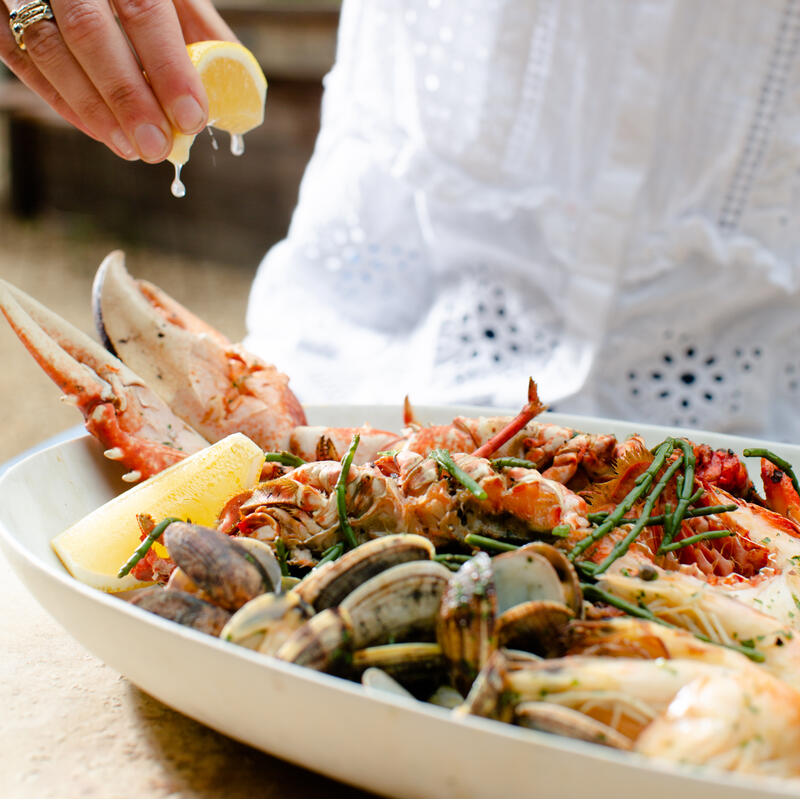 A person squeezes lemon juice onto a seafood platter with crabs, clams, shrimp, and greenery, set outdoors.