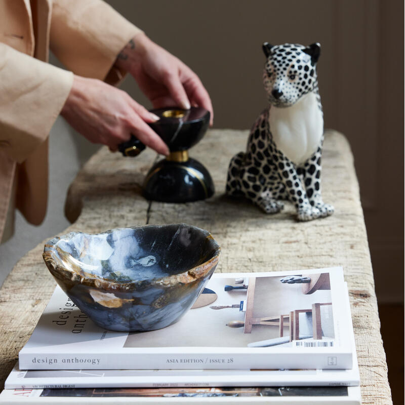 Ceramic bowl resting on stacked magazines sits atop a rustic wooden table. Nearby, a person in a beige suit adjusts a black and gold object beside a spotted ceramic leopard. Text includes "design anthology ASIA EDITION / ISSUE 28" and dates of various "AR