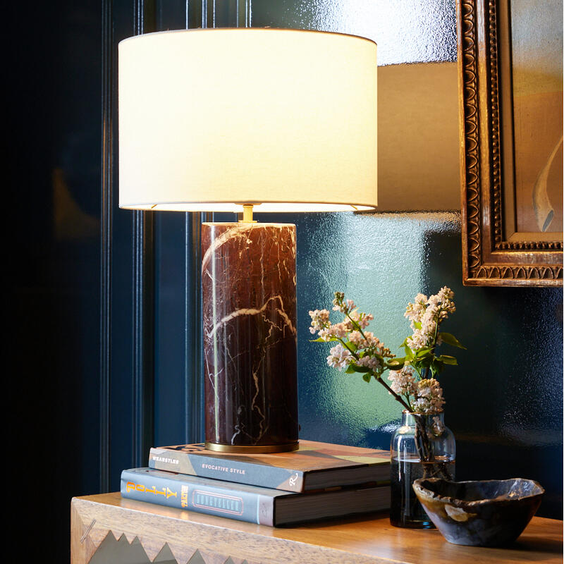 A lamp with a white shade on a wooden table, next to a stack of books, a vase of flowers, and a ceramic bowl, in a room with dark blue walls and framed art. The book titles are "Wearstler," "Evocative Style," and "The 3D Type Book."