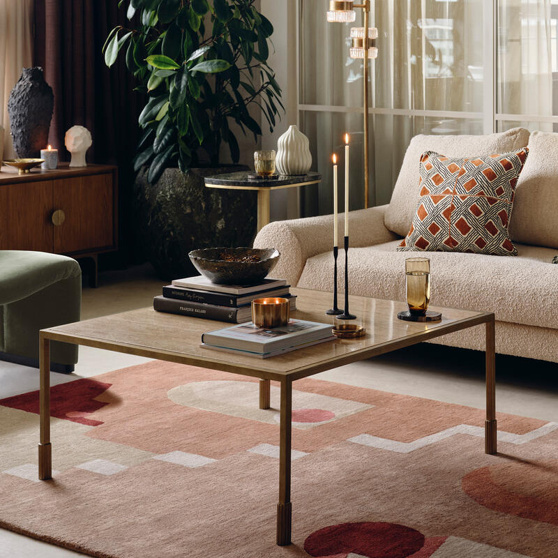 A beige couch with patterned cushions stands in a modern living room, surrounded by green plants, a coffee table with candles and books, an armchair, and large windows with sheer curtains.