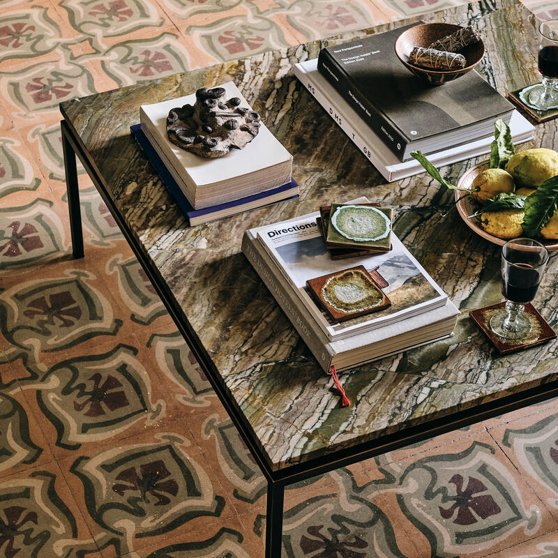 A marble-top table holds books, a bowl of lemons, green coasters, wine glasses, and a decorative object, set on ornate patterned floor tiles in a cozy indoor setting. The visible book titles are “Directions: “Exploration sketchbook black and white photo r