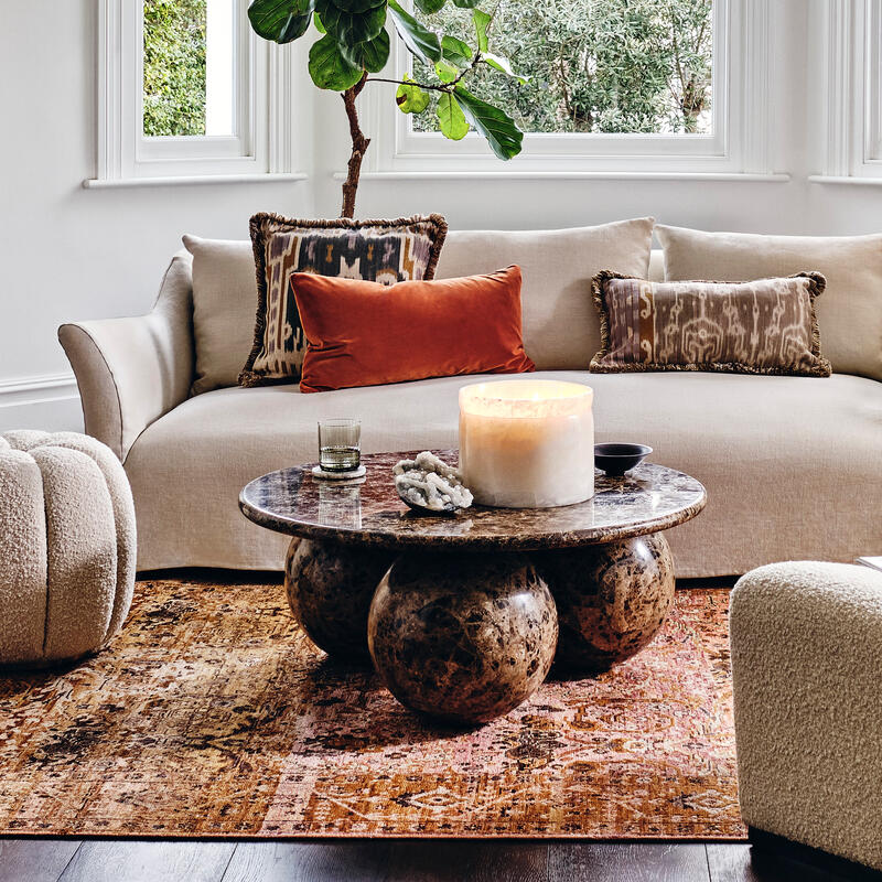 A beige sofa with decorative pillows stands in a stylish living room, surrounded by a large window, indoor plant, round ottoman, textured chairs, and a coffee table on a patterned rug.