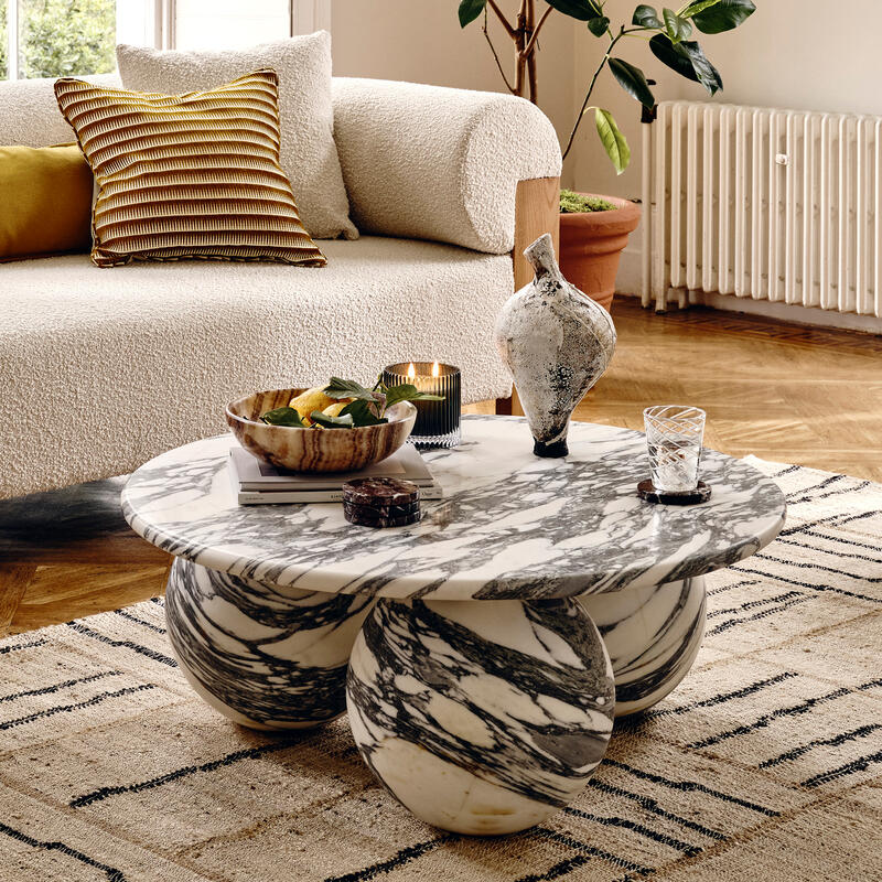 A living room features a white textured sofa, a marble coffee table with spherical legs holding decor, and a large potted plant. A circular, amber chandelier hangs above, near tall windows.