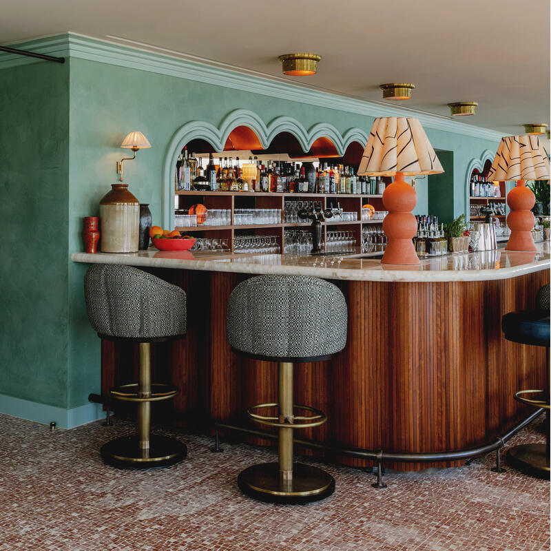 A curved wooden bar with marble top and patterned lamps, flanked by cushioned bar stools, is set against a soft green wall with a mirror and shelf of alcohol bottles.