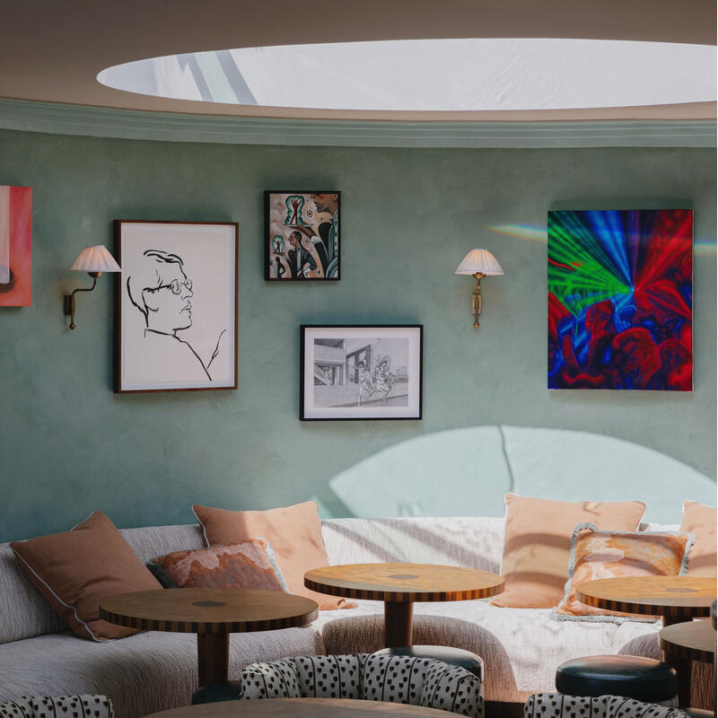 A cozy seating area features a curved sofa with cushions, surrounded by round wooden tables and chairs. The mint-green wall displays various artworks, illuminated by wall lamps and natural light from a skylight.