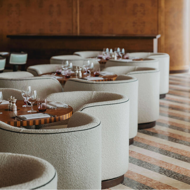 Curved, beige chairs surround wooden tables set with glasses and cutlery in a sophisticated restaurant with a patterned tile floor and wooden walls.