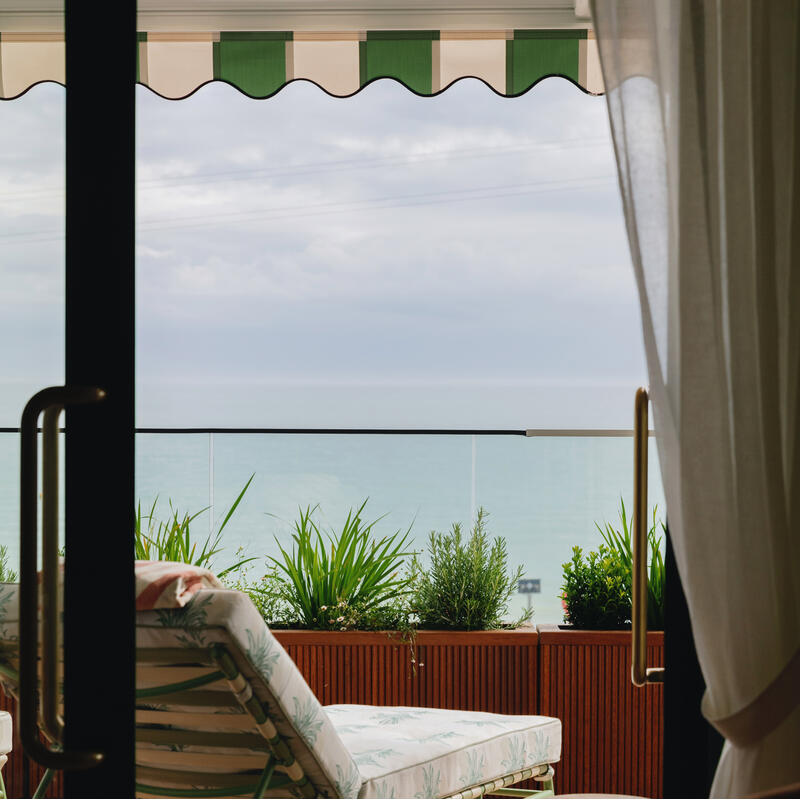 A cushioned lounge chair faces an ocean view on a balcony with potted plants, under a green and white striped awning, viewed through a partially open sliding glass door with white curtains.