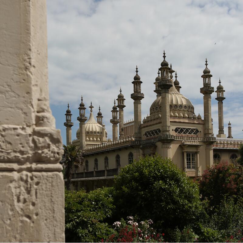 A grand, ornate building with multiple white domes and minaret-like spires stands amidst lush greenery and blooming flowers under a partly cloudy sky, partially obscured by a textured stone structure.