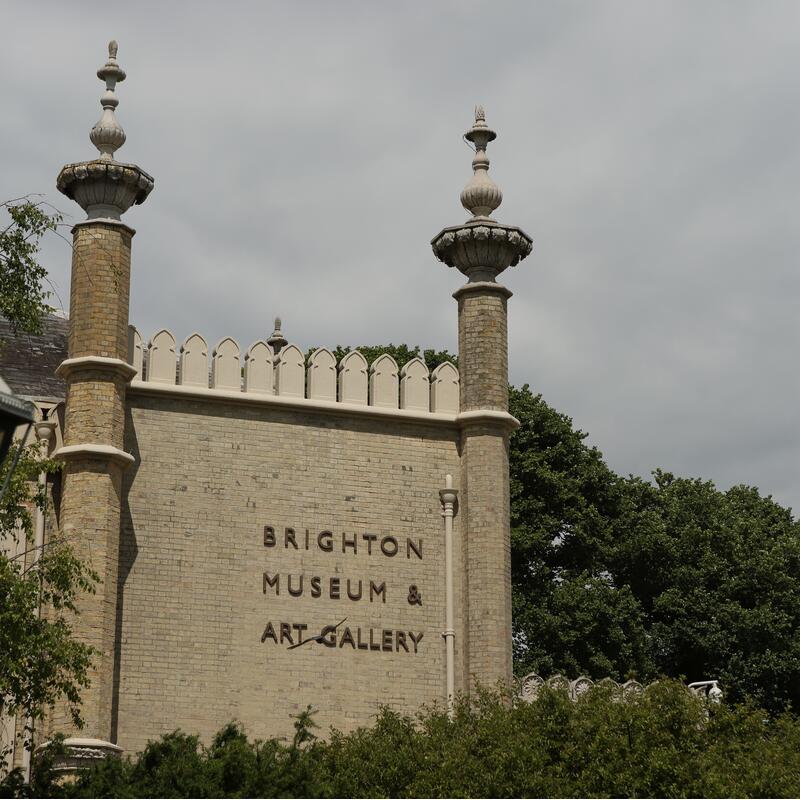 A brick building labeled "BRIGHTON MUSEUM & ART GALLERY" features ornate towers. It is surrounded by greenery and set against a cloudy sky.