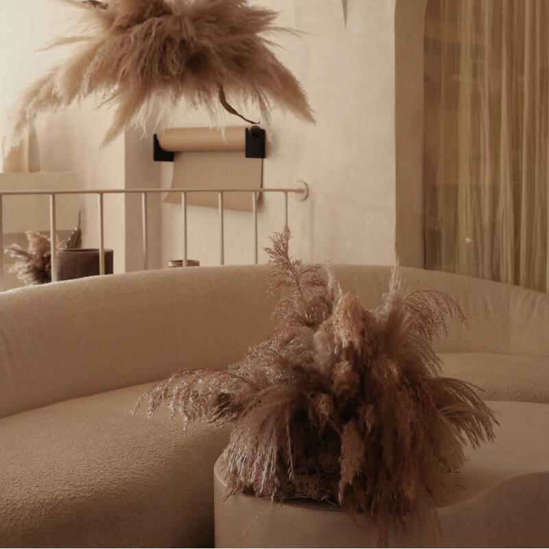 A sculptural beige couch adorned with fluffy pampas grass is placed in a minimalist living room containing a light beige wall and a textured accent wall.