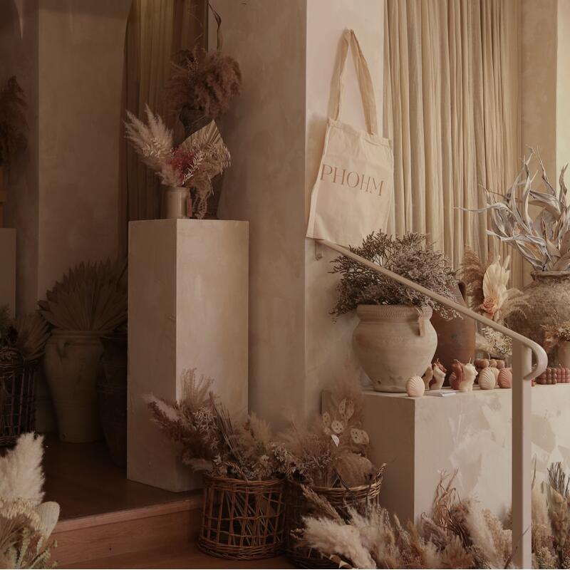 Baskets filled with dried flowers rest on wooden stairs in a warm-toned room adorned with rustic vases and a tote bag that reads "PHOHM", hanging beside a curtained wall.