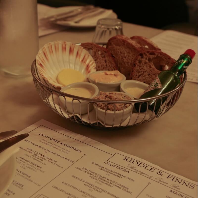 A wire basket with assorted bread, sauces, and a Tabasco bottle on a table, surrounded by water glasses, cutlery on a plate with a napkin, and a menu from "RIDDLE & FINNS."