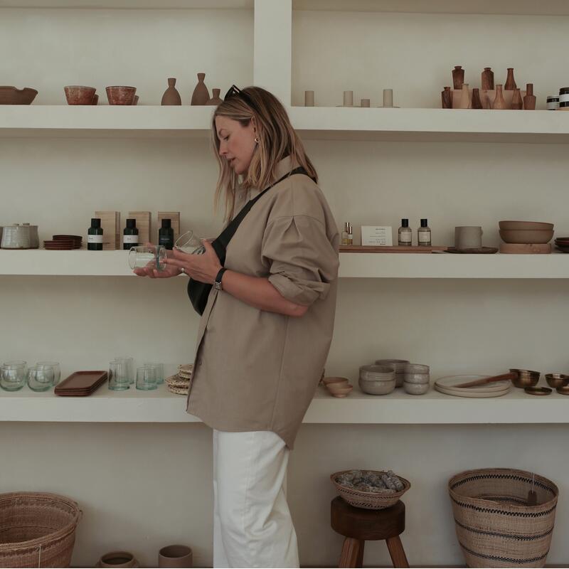 A woman carefully examines glassware in a minimalist shop featuring white shelves filled with various home decor items, including bowls, jars, and woven baskets.