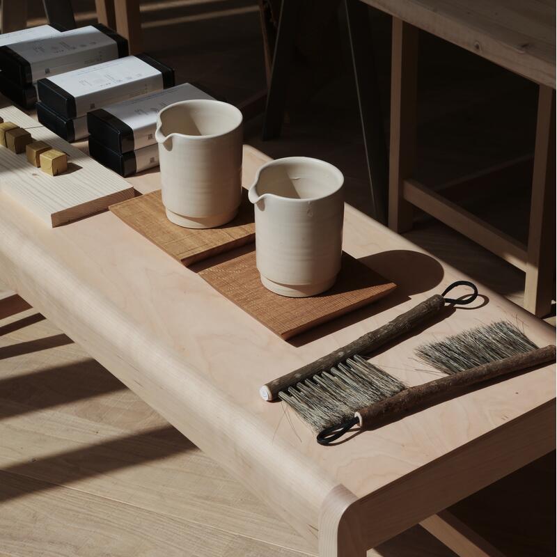 Ceramic pitchers rest atop wooden placemats on a light wood table, which also holds brushes with worn bristles and packaged goods under soft sunlight in a rustic, minimalistic setting.