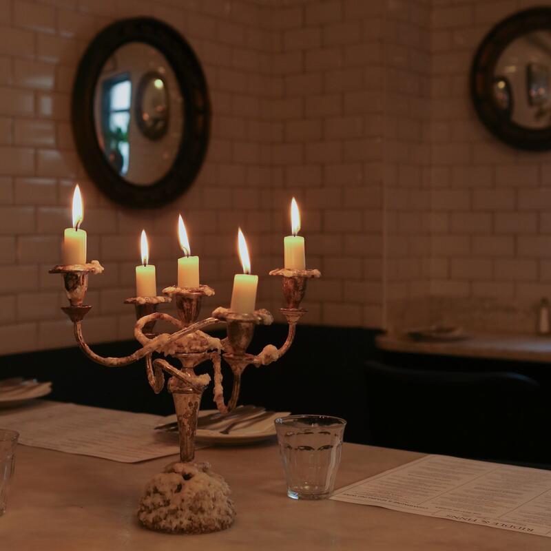 Candlestick holder with lit candles on a restaurant table, surrounded by glass, silverware, plates, and menus, against a backdrop of white tiled walls and round mirrors.