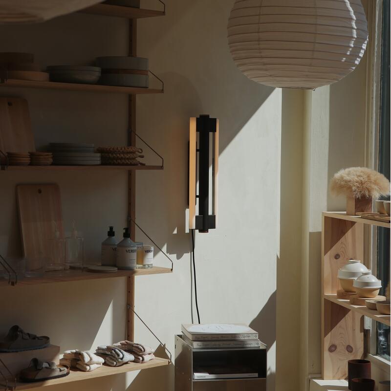 Household items sit neatly arranged on wooden shelves in a well-lit corner shop, with sunlight streaming through a large window. A modern wall lamp provides additional ambient light.