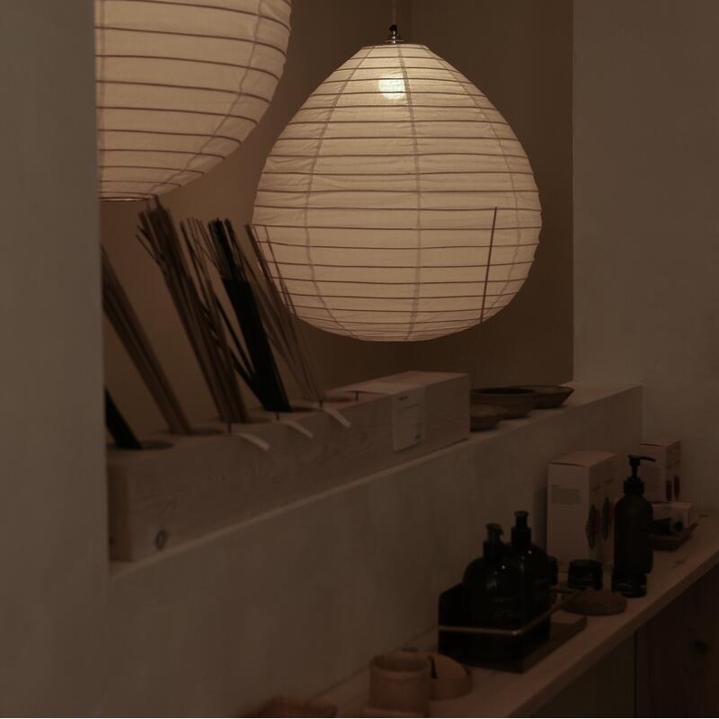 Two paper lanterns hang, softly glowing. Below them, incense sticks and bottles are arranged on wooden shelves. The setting is a dimly lit, minimalist room.