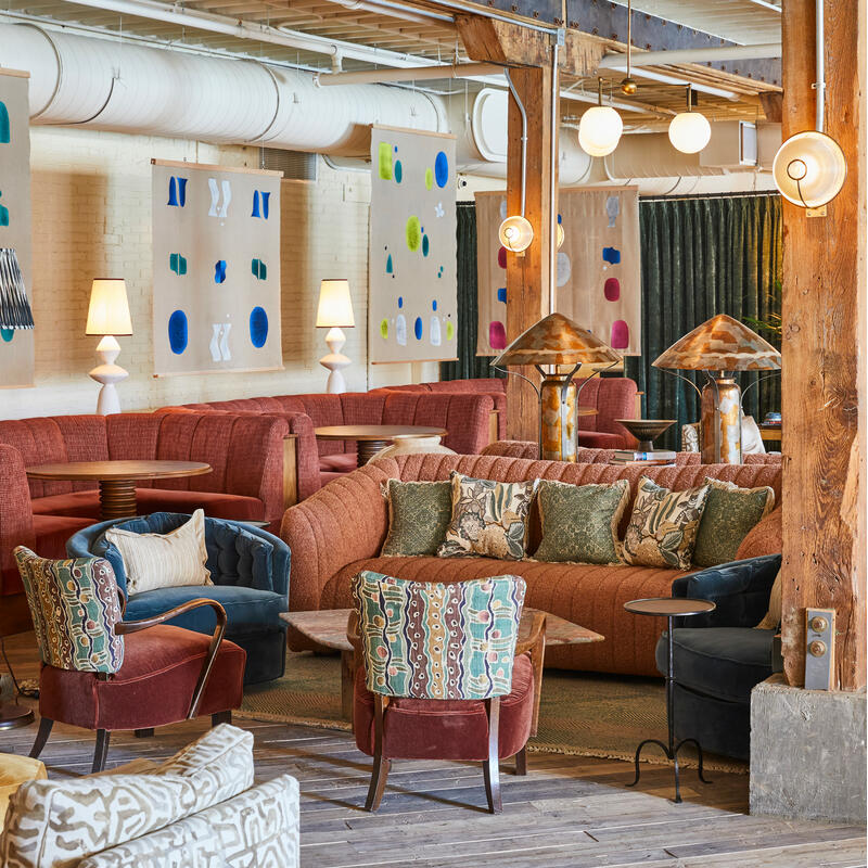 An eclectic lounge area with colorful couches and armchairs, decorated with abstract wall art and lamps, supported by wooden beams, set in a spacious, industrial-chic environment.