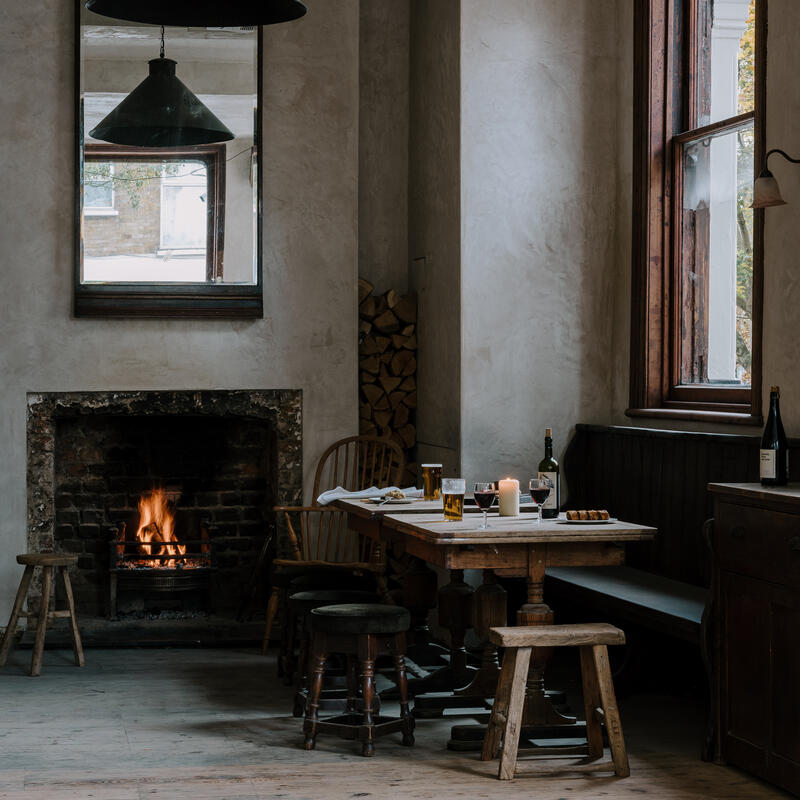 A cozy fireplace with a burning fire next to a wooden dining table with drinks and food, in a rustic room with a large window and a mirror. Text on the mirror reads "DISPENSING DEPARTMENT."