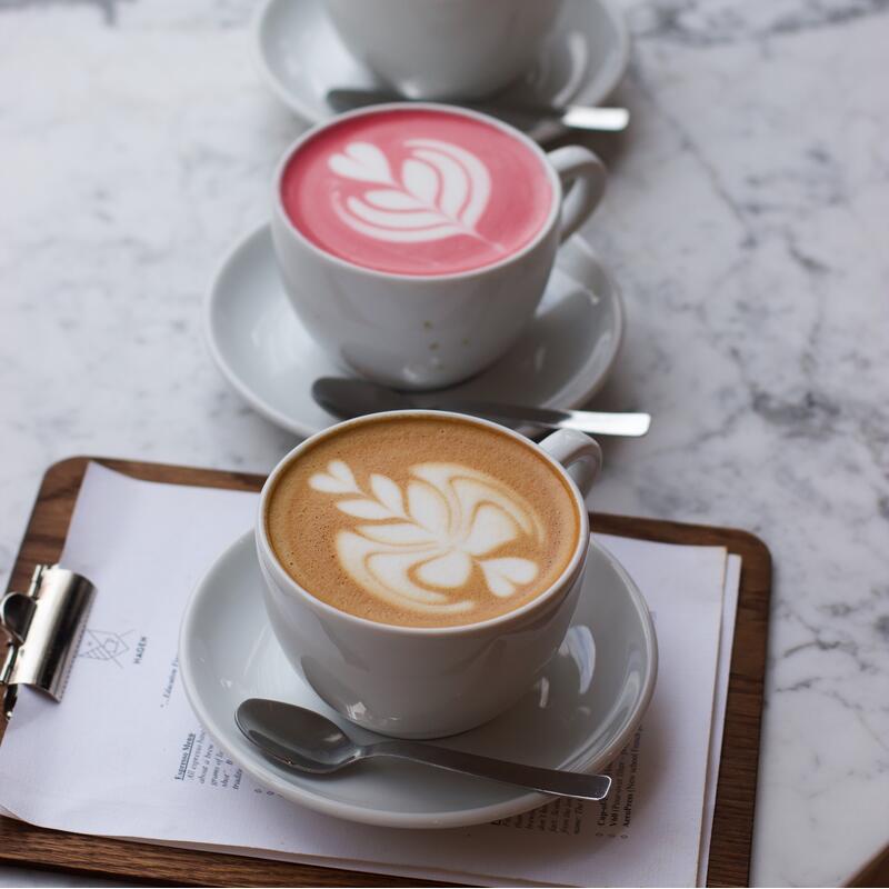 Three cups of lattes with intricate foam art, featuring black, pink, and beige colors, respectively, are arranged on white saucers on a marble table with a clipboard beneath.