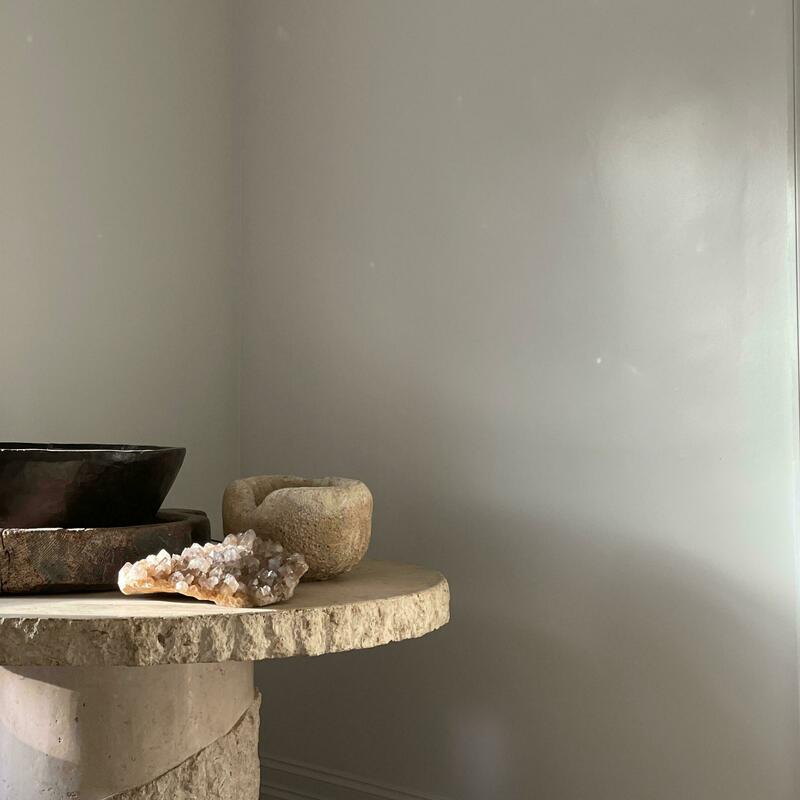 A large stone bowl, a small textured bowl, and a geode crystal rest on a round, rough stone table in a minimally decorated room with light gray walls.