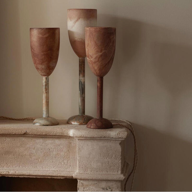 Three marble goblets, in varying shades, stand atop a rough, beige stone mantel against a plain wall. Coils of rope lie beside the goblets, adding to the minimalistic ambiance.