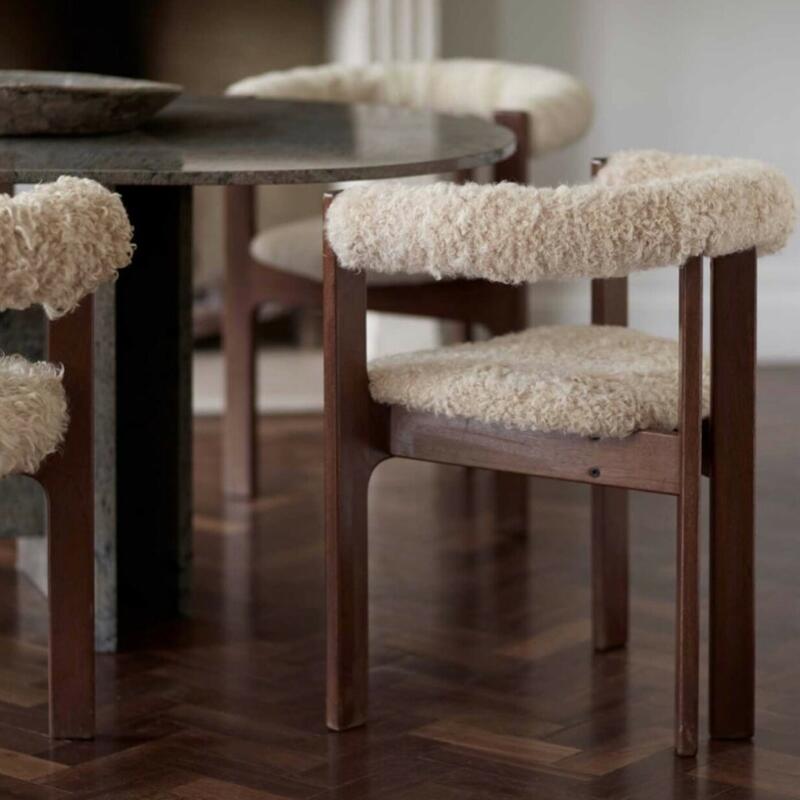 A wood-framed chair with plush, white, curly upholstery positioned around a round, stone-top table in a room with parquet flooring and neutral wall colors.