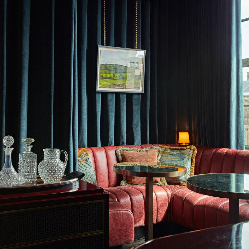 A crystal decanter set sits on a tray atop a wooden cabinet in a luxurious room with red velvet couches, round tables, and a large window revealing historical architecture outside.