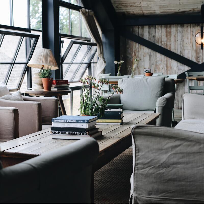 Books and a potted plant sit on a wooden coffee table surrounded by armchairs in a cozy, rustic loft with large windows and wooden walls. Text on books: "The Principles of Architecture (Foster)", "People Rofoff Beny".