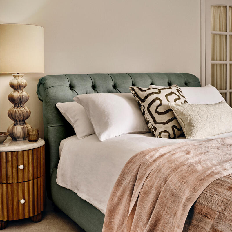 Bed adorned with white linens and patterned pillows. Beside it, a wooden nightstand with a sculpted lamp. Situated in a cozy, neutrally colored bedroom.