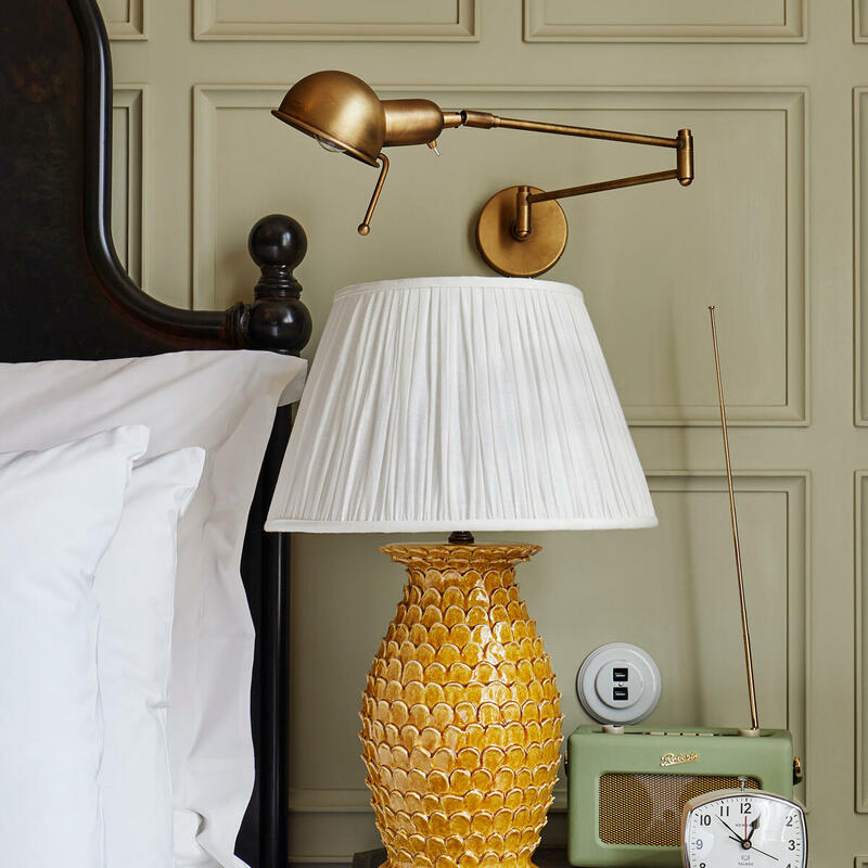 A yellow textured lamp with a white shade stands on a wooden nightstand with an alarm clock and vintage radio, next to a bed with white linens and a brass wall-mounted reading light.