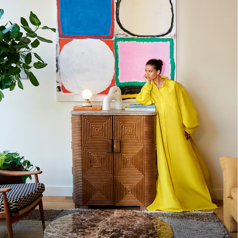 A woman in a yellow dress leans on a wooden textured cabinet under a bright chandelier. Behind her is a colorful abstract painting, and a marble table and plants are visible in the room.