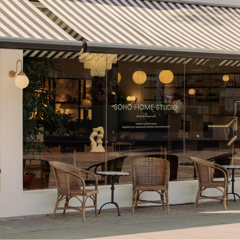 Wicker chairs and small tables are arranged outside a storefront with large windows and striped awnings. Text on the window reads: "SOHO HOME STUDIO, inspired by Soho House interiors."