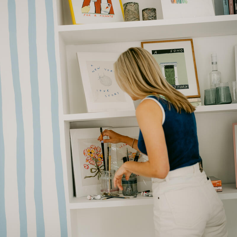 A woman in a blue top and white pants organizes art supplies on a white shelf, with framed artwork and various items around her in a striped-wall room. "A NEW YORK LOVE STORY" is text written on one of the artworks.