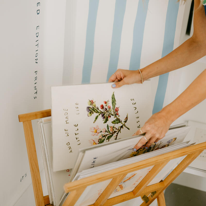 Hands sorting through limited edition prints in a wooden stand, with blue striped walls and framed art; some prints depict flowers with text reading "AND WE MADE LITTLE LIVES LIKE OURSELVES."