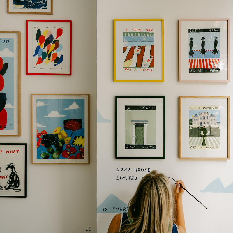 A person paints on a wall adorned with framed, colorful illustrations featuring texts like "HAVE SOME FUN," "LET'S HAVE SOME FUN," and "IT'S A WONDERFUL DAY IN SOMERSET," in a bright room. Text on wall reads: "SOHO HOUSE LIMITED IS THERE."