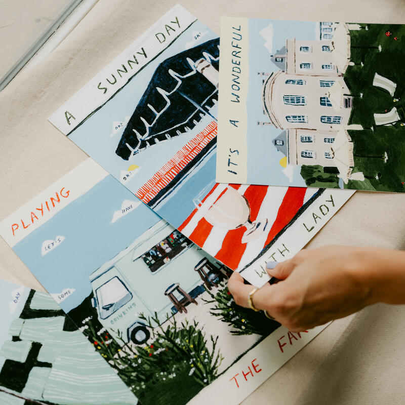 Several colorful illustrated postcards lie on a beige surface, with a hand picking one up. Text readable: "A SUNNY DAY", "IT’S A WONDERFUL", "PLAYING WITH LADY", "ON THE FARM."