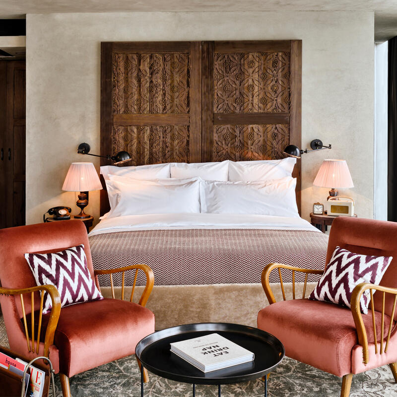 A well-appointed bedroom features a large bed with ornate wooden headboard, twin bedside lamps, and two upholstered chairs with patterned cushions, set within a contemporary-styled room with wooden accents.