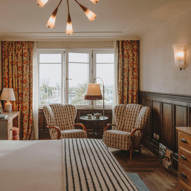 A stylish bedroom features a large bed with patterned pillows, a modern chandelier, two armchairs near a window with floral curtains, wooden furniture, and decorative lighting, creating an elegant and cozy ambiance.