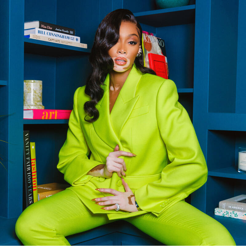 A person wearing a bright green suit sits confidently within a blue bookshelf setting. Books, candles, and vases are arranged on the shelves. 

Visible book titles: 
- "Miss Dior" by Justine Picardie
- "Captivate" by Vanessa Van Edwards
- "Kimono" edited 