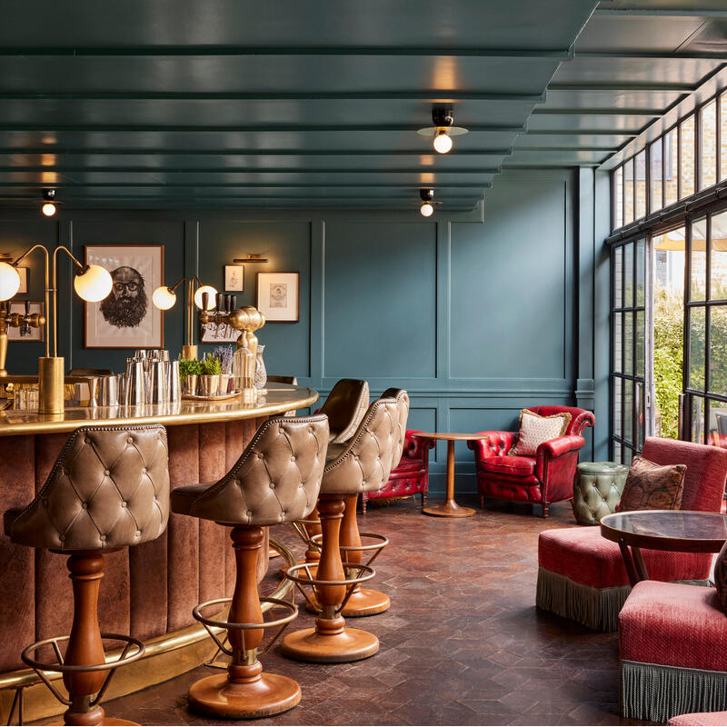 Bar area with leather stools at a counter featuring lamps, cocktail shakers, and greenery. Surrounding are upholstered red chairs and tables in a warmly lit, paneled room with large windows.