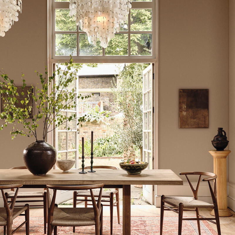 Wooden dining table set with decor under two chandeliers, framed by large windows and open doors, revealing a lush garden outside. Accessories include a vase, candles, and a fruit bowl.