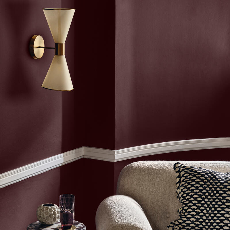 Beige wall lamp is mounted above a marble side table with a candle holder and glass, next to a beige sofa with a patterned cushion set in a room with dark red walls.