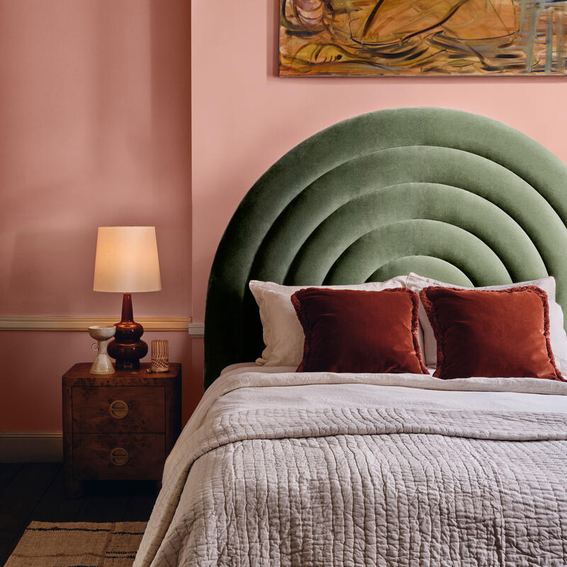 A bed with a curved green headboard and white quilted bedding, flanked by a wooden nightstand with a lamp, set against a pink wall with an abstract painting.